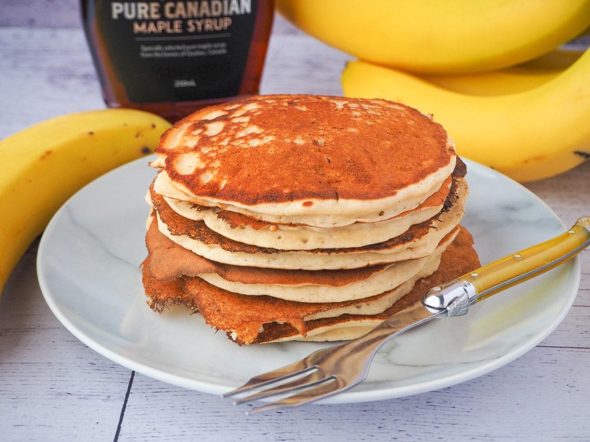 Stack of banana pikelets on a plate, with a vintage spoon on the side, surrounded by fresh whole bananas and bottle of maple syrup in the background.