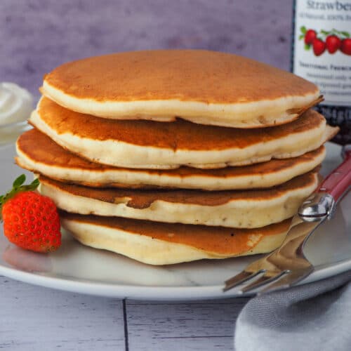 Close up stack of pikelets on a plate, with a fresh strawberry and fork, and whipped cream and jar of strawberry jam in the background,