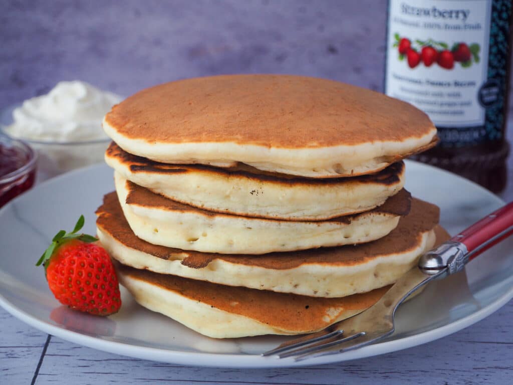 stack of pikelets on a plate, with a fresh strawberry and for, and whipped cream and jar of strawberry jam in the background,
