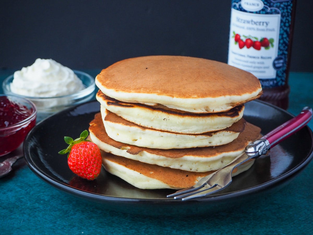 stack of pikelets on a plate, with a fresh strawberry and for, and whipped cream and jar of strawberry jam in the background,