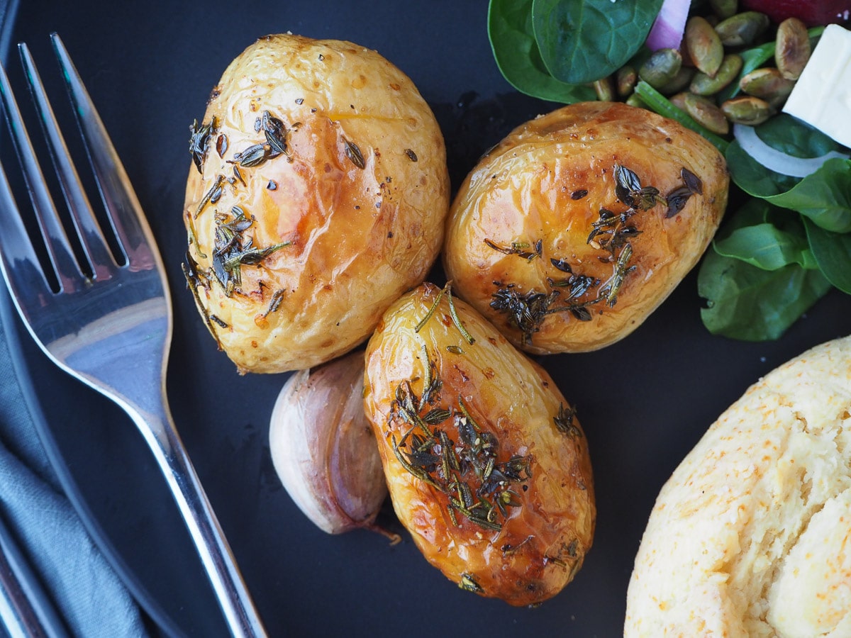 Close up of roasted mini potatoes on a black plate, with beetroot salad and damper on the side