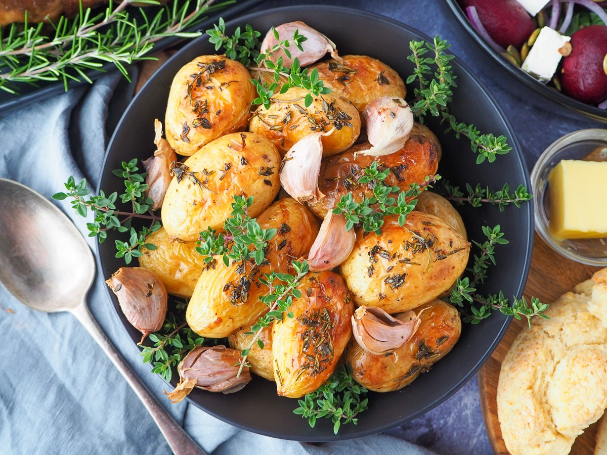 Roasted mini potatoes in a serving dish garnished with fresh herbs, silver serving spoon to the side, fresh damper, pat of butter, beetroot salad and slow cooker roast lamb on the side.