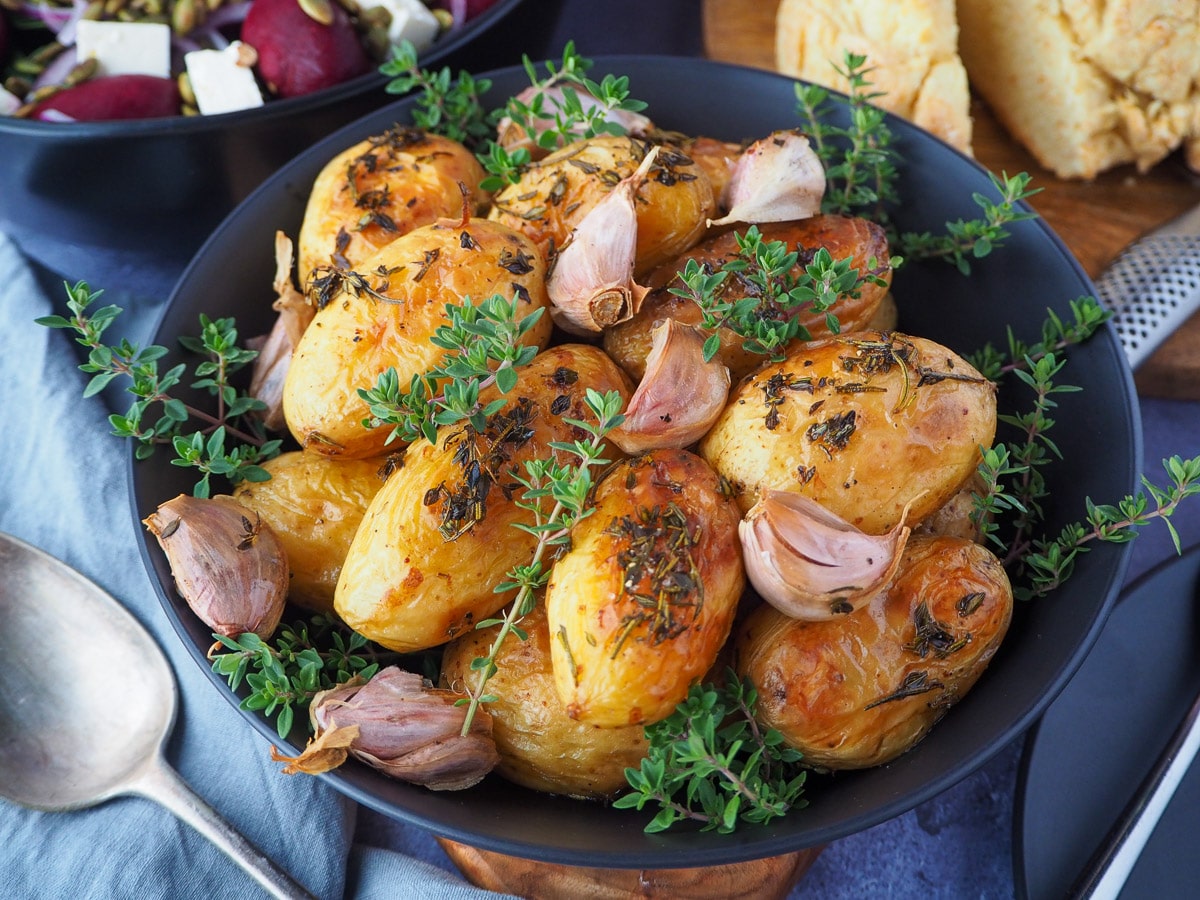 Roasted mini potatoes in a serving dish garnished with fresh herbs, silver serving spoon to the side, fresh damper, pat of butter, beetroot salad and slow cooker roast lamb on the side.