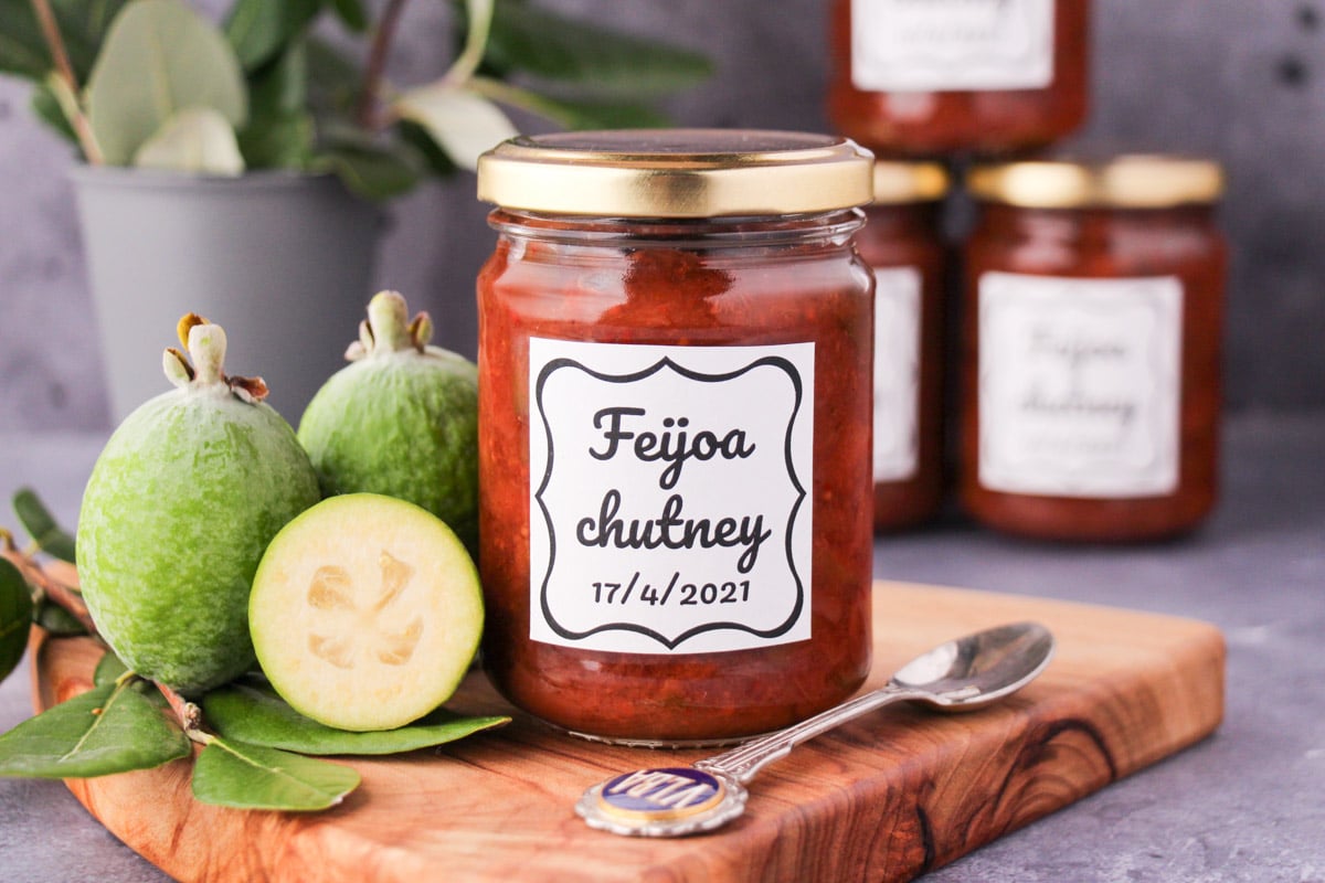 jar of homemade feijoa chutney on a board, with vintage spoon and fresh feijoas, stacked jars of chutney in the background.