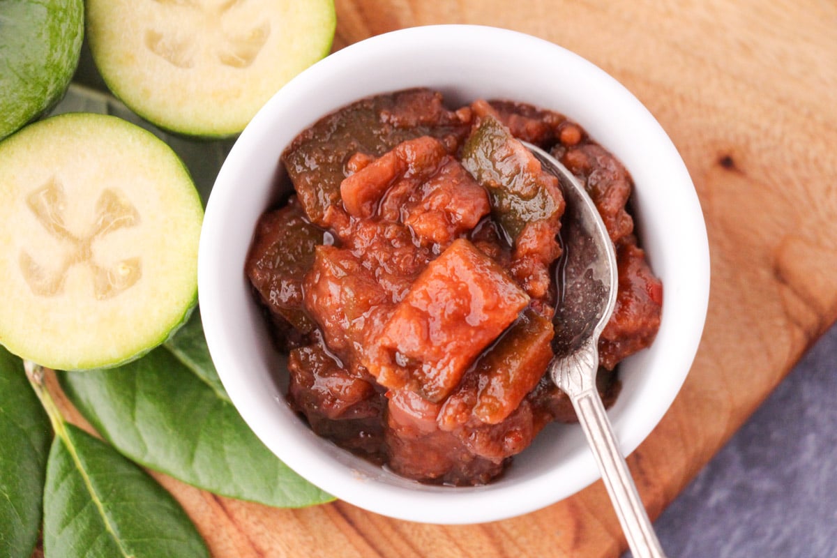 Feijoa chutney in a serving bowl on a bowl with a serving spoon, on a board with fresh feijoas and feijoa leaves.