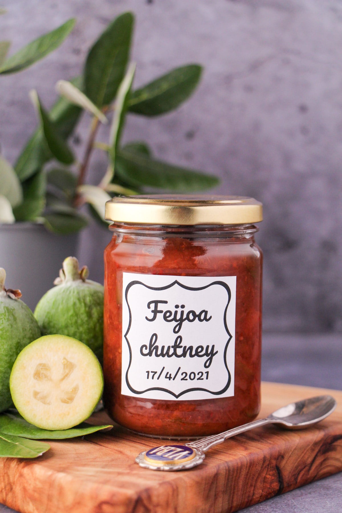 Jar of homemade feijoa chutney on a board, with vintage spoon and fresh feijoas, and fresh feijoa leaves in the background.