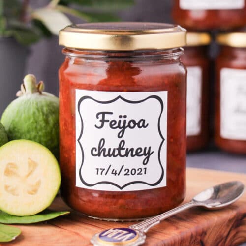 Close up jar of homemade feijoa chutney on a board, with vintage spoon and fresh feijoas, stacked jars of chutney in the background.