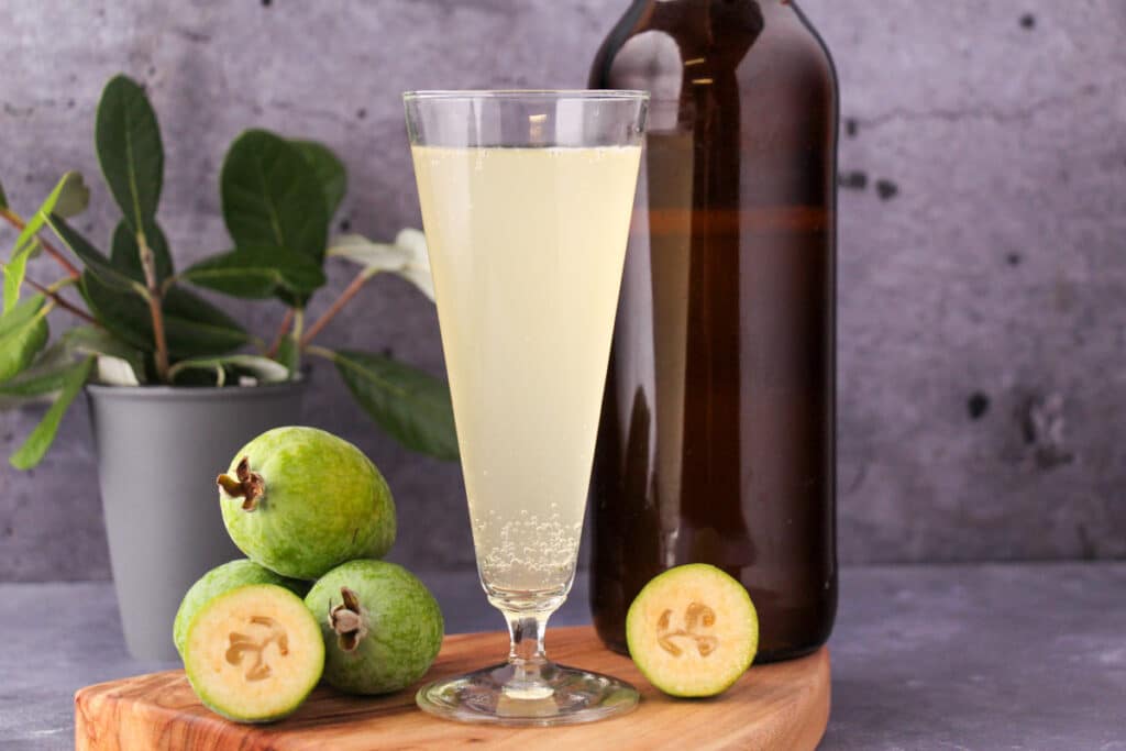 Tall glass of feijoa fizz on a board, with bottle of feijoa fizz, fresh feijoas and feijoa leaves.