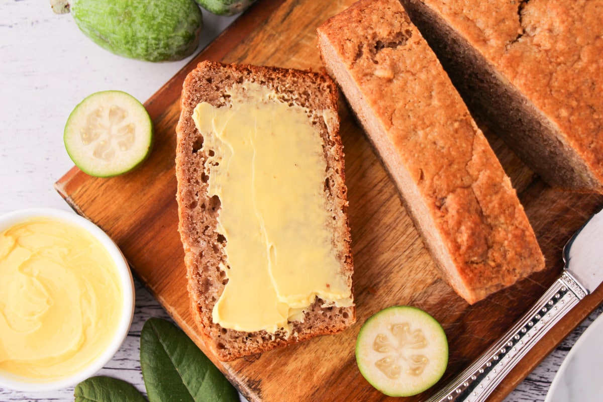Sliced feijoa loaf on a chopping board, with one buttered slice, fresh feijoas and leaves and a butter knife.