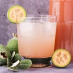 Close up glass of made up feijoa syrup with ice and fresh feijoa garnish, with bottle of feijoa syrup and fresh feijoas and leaves.