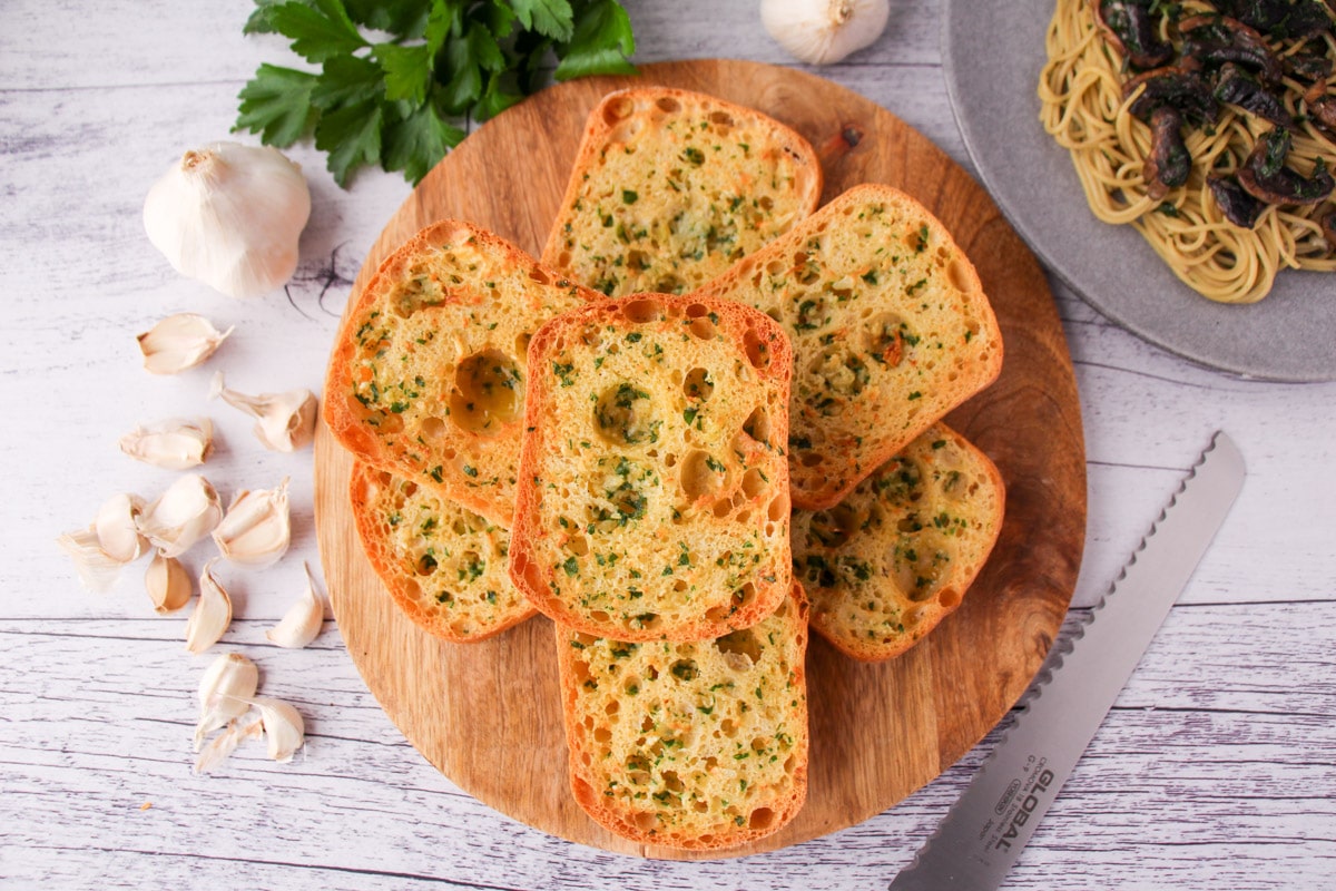 Stack of air fryer garlic bread on a chopping board, with bread knife, fresh garlic cloves, fresh parsley and a plate of pasta on the sides.
