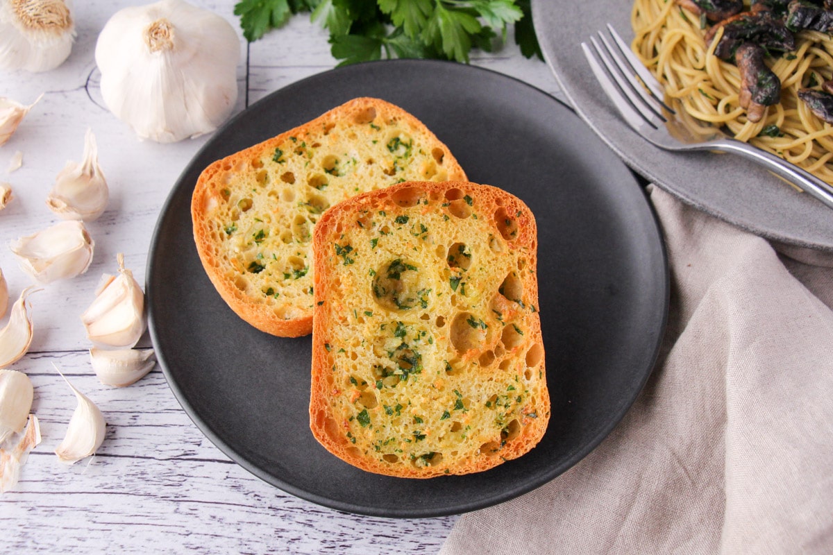 Air fryer garlic bread on a plate, with fresh garlic cloves, fresh parsley and a plate of pasta on the sides.