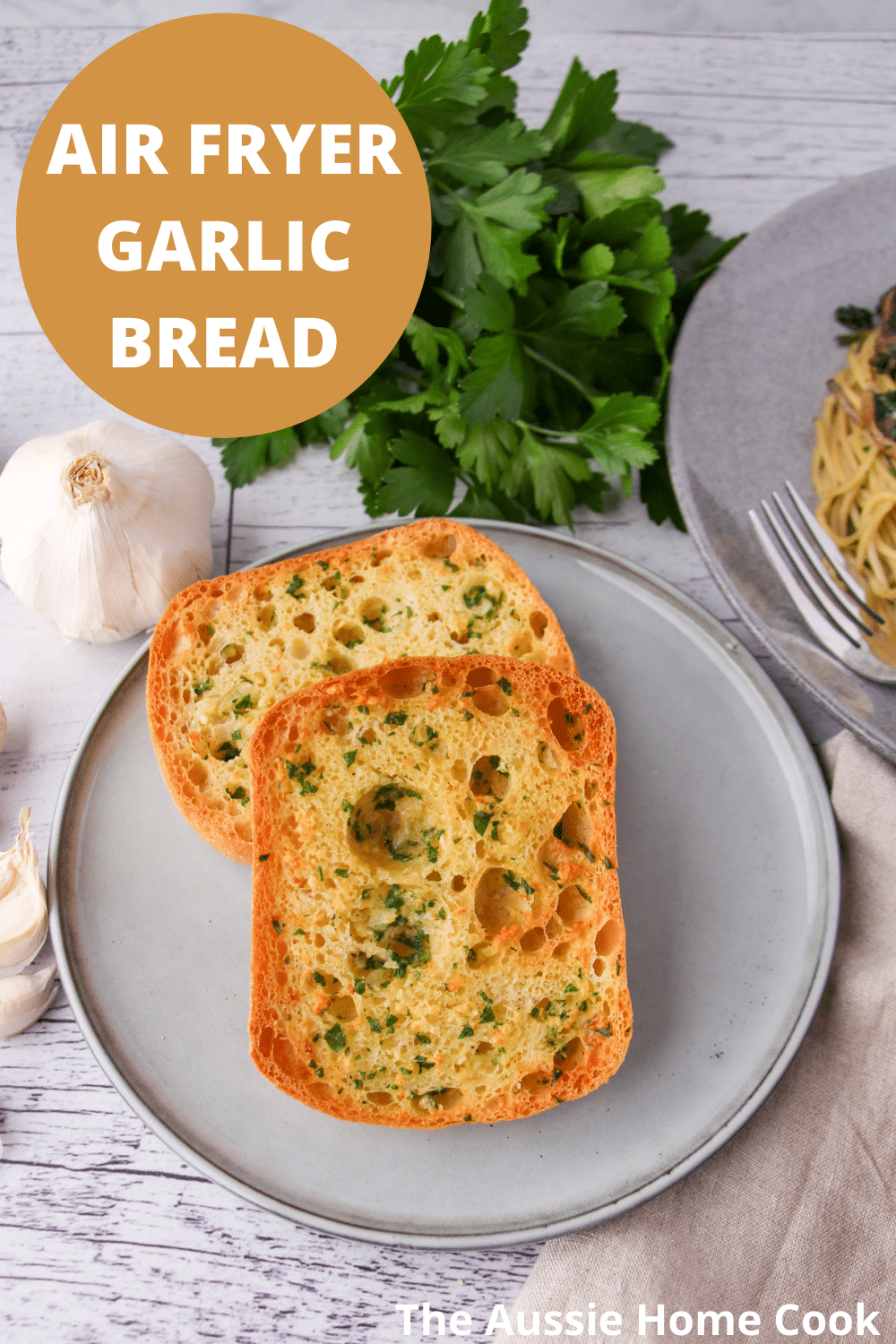 Air fryer garlic bread on a plate, with fresh garlic cloves, fresh parsley and a plate of pasta on the sides, with text overlay, Air fryer garlic bread and The Aussie Home Cook.