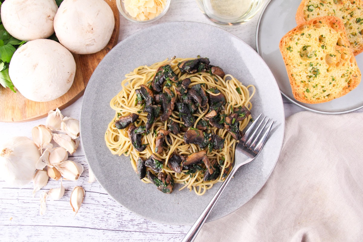 Mushroom aglio olio on a plate with a fork, with garlic bread, fresh garlic, mushrooms and parsley, a glass of white wine and shaved parmesan around the edges.