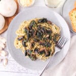 Close up mushroom aglio olio on a plate with a fork, with garlic bread, fresh garlic, mushrooms and parsley, a glass of white wine and shaved parmesan around the edges.