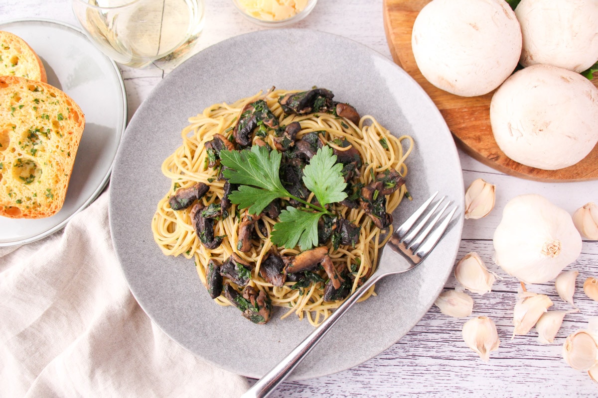 Mushroom aglio olio on a plate with a fork, with garlic bread, fresh garlic, mushrooms and parsley, a glass of white wine and shaved parmesan around the edges.