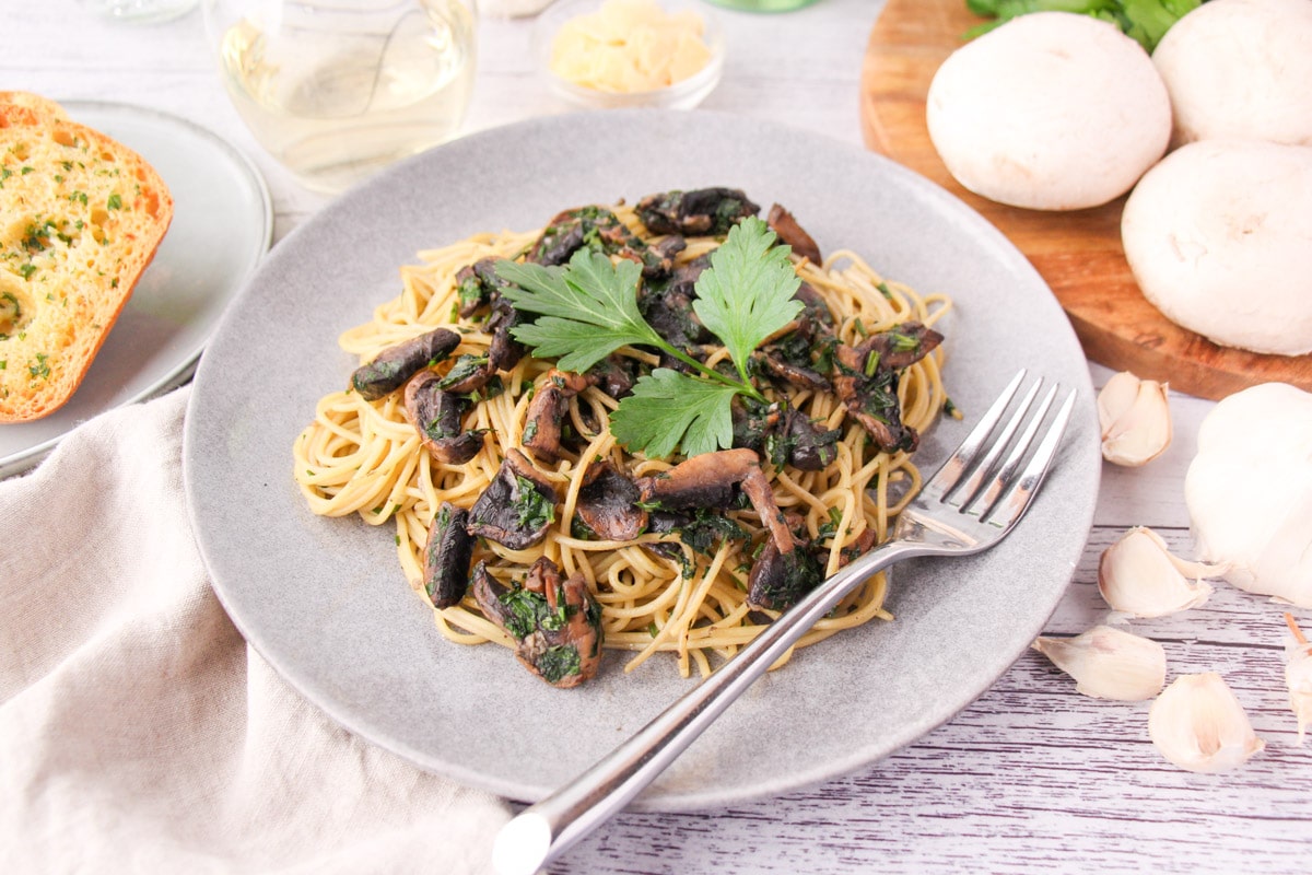 Mushroom aglio olio on a plate with a fork, with garlic bread, fresh garlic, mushrooms and parsley, a glass of white wine and shaved parmesan around the edges.