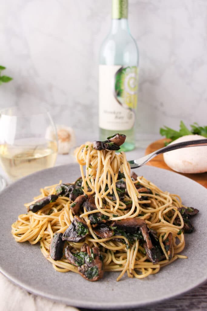 Fork of mushroom aglio olio held over a plate of pasta, with fresh mushrooms, parsley, garlic and a glass and bottle of white wine the background.