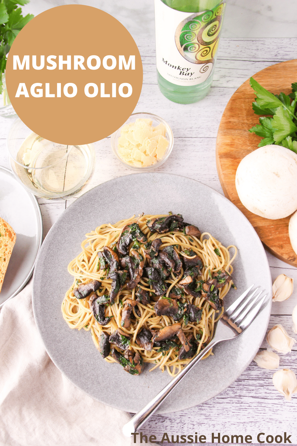 Mushroom aglio olio on a plate with a fork, with garlic bread, fresh garlic, mushrooms and parsley, a glass and bottle of white wine and shaved parmesan around the edges., with text overlay, mushroom aglio olio, The Aussie Home Cook.