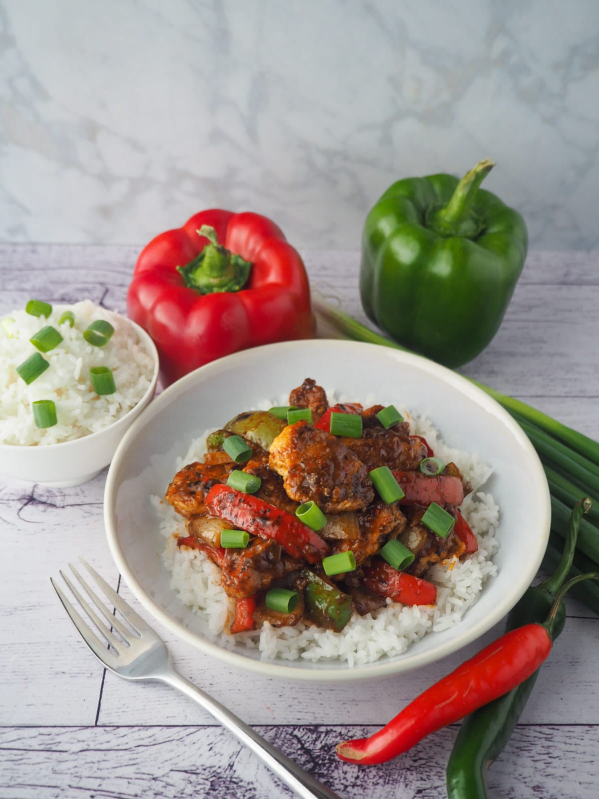 Bowl of chicken on rice with fork, bowl of rice, capsicums, spring onions and chilli on the side.