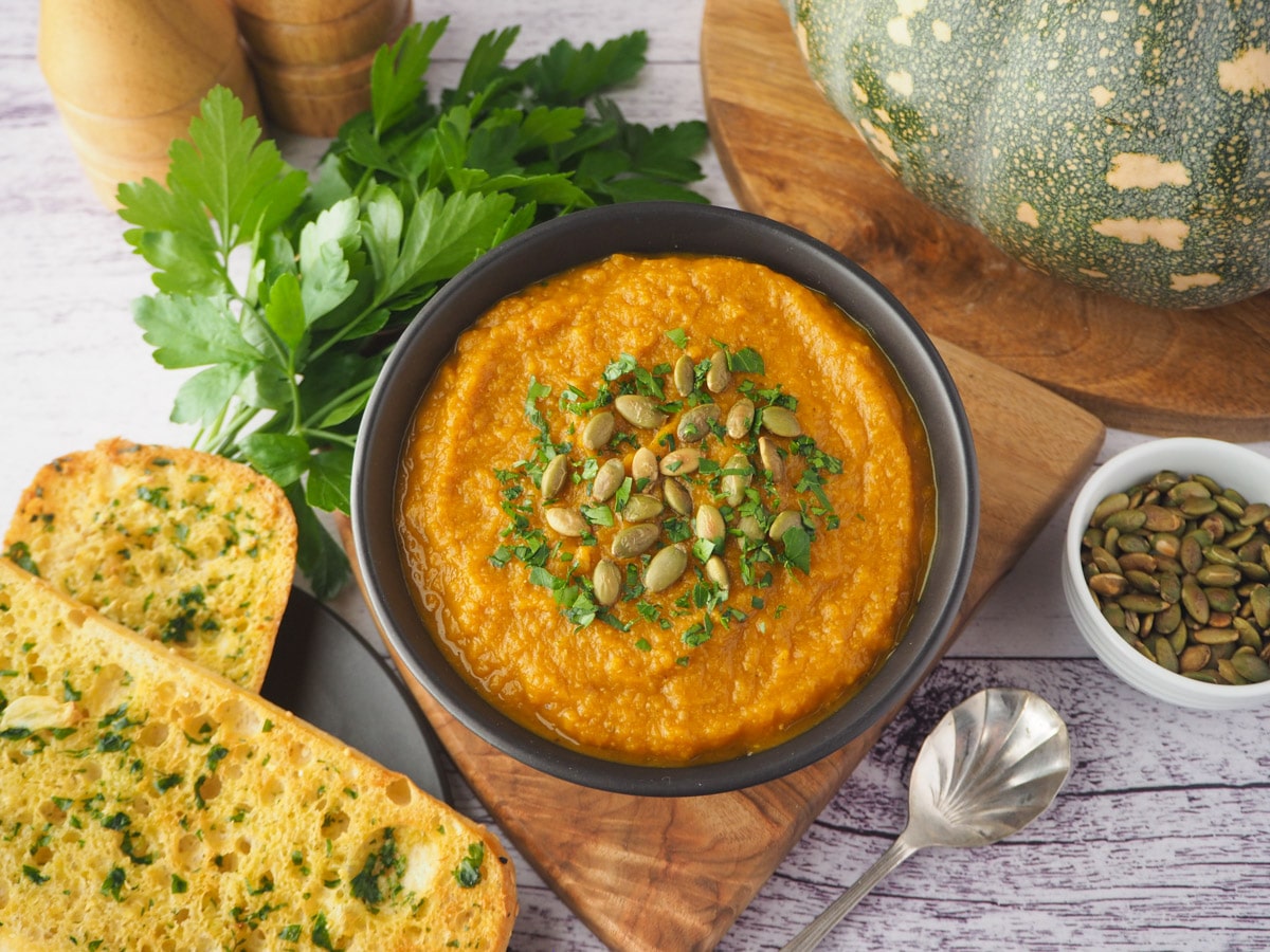 Soup with pumpkin seed and parsley garnish, with garlic bread, spoon and toasted pumpkin seeds on the side.