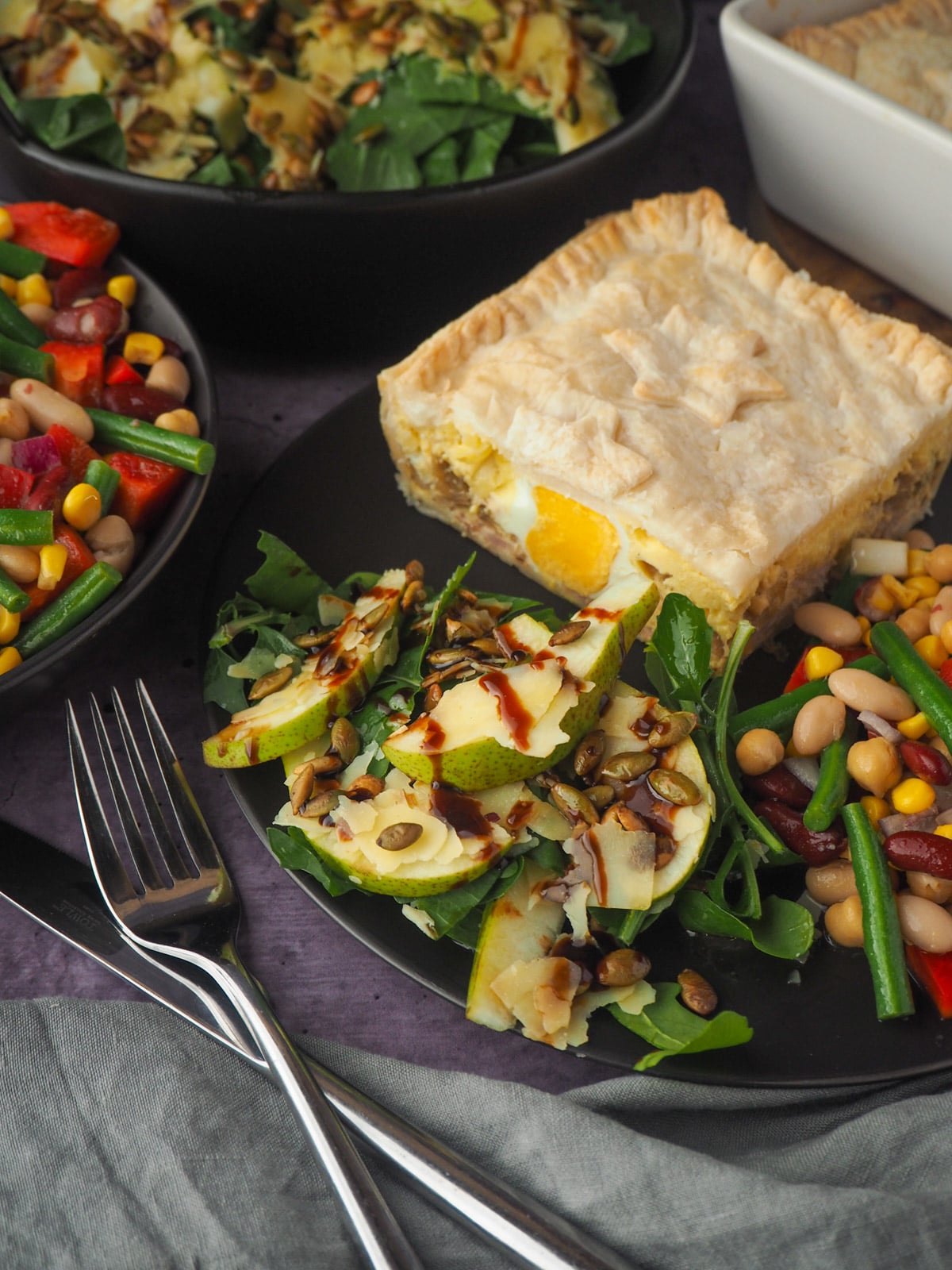 Serving of salad on a plate with bacon and egg pie and four bean salad, knife and fork to the side and salads and pie in the background.