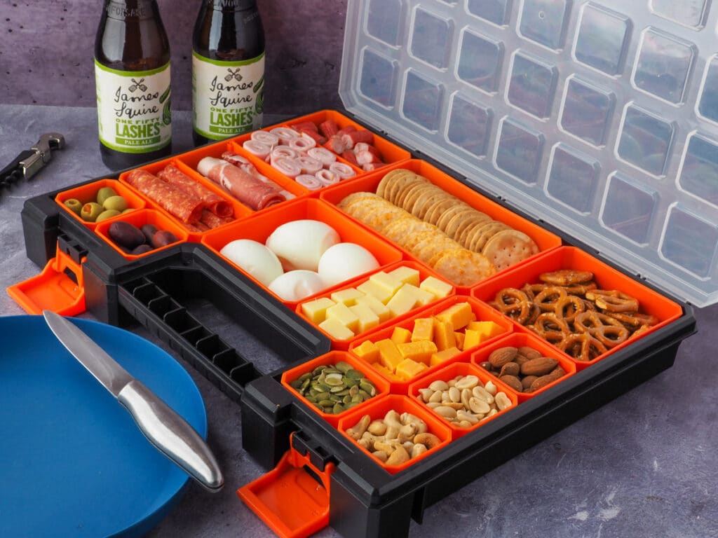 Multi compartment container filled with various charcuterie food, with picnic plate and drinks in the background.