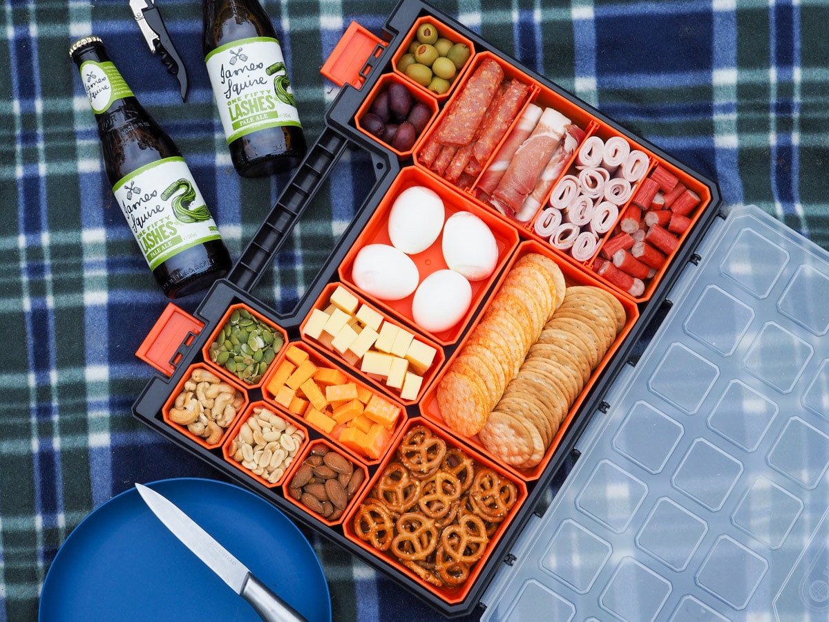 Multi compartment container filled with various charcuterie food, on a picnic blanket with a plate and drinks.