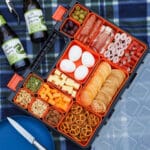 Multi compartment container filled with various charcuterie food, on a picnic blanket with a plate and drinks.