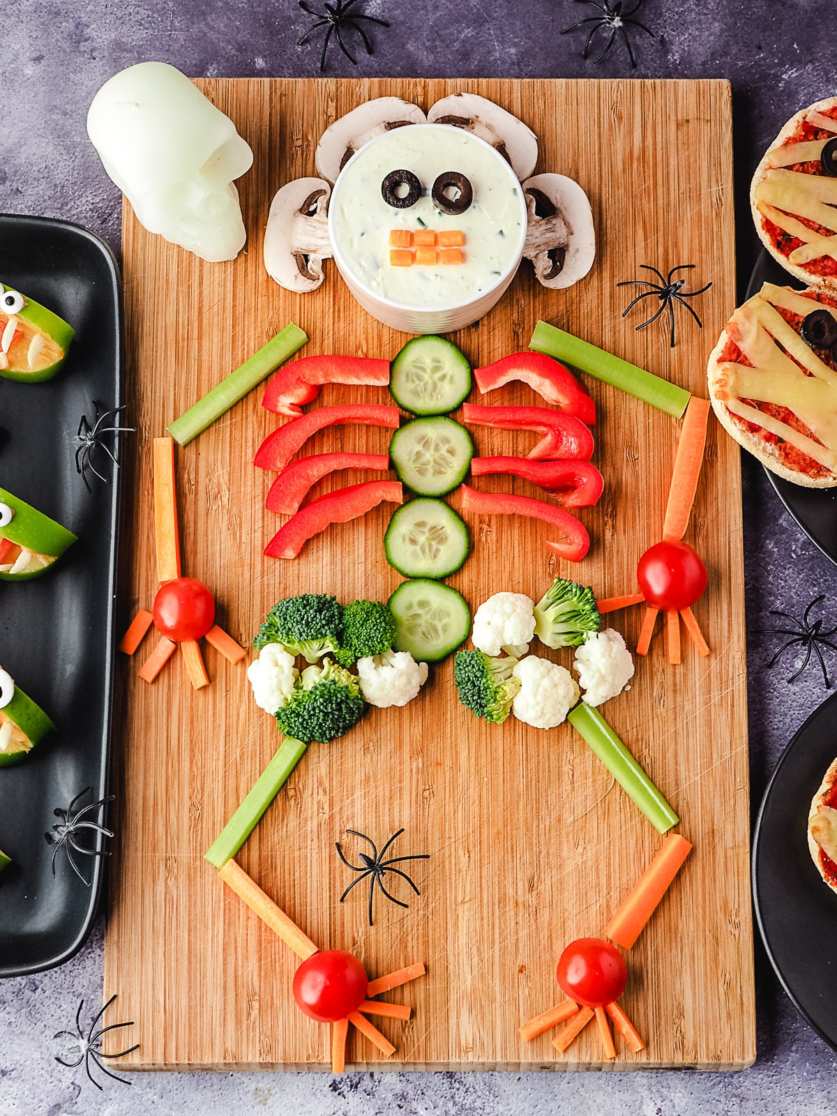 Veggie tray on a board, with halloween skull and spiders, surrounded by Halloween party food, apple monsters and mummy pizzas.