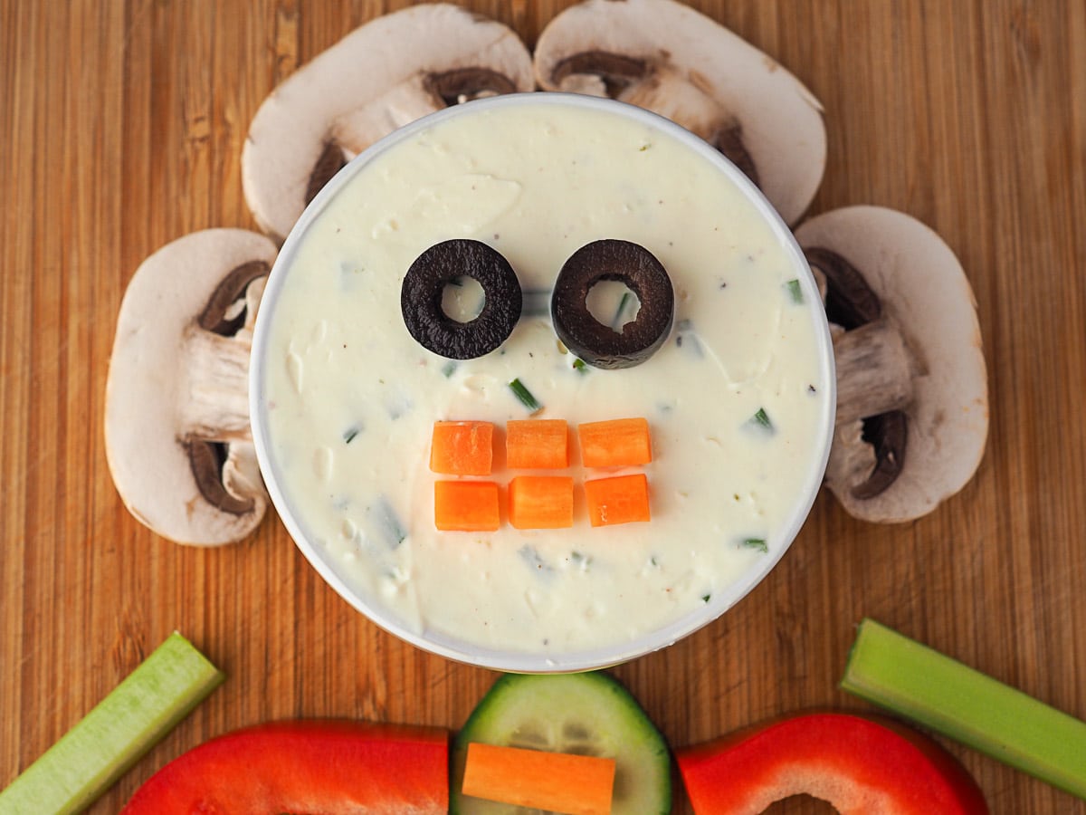 Process shots: dip showing two sliced olive pieces as eyes and carrot cut into teeth shapes. Dip is surrounded by mushroom slices.