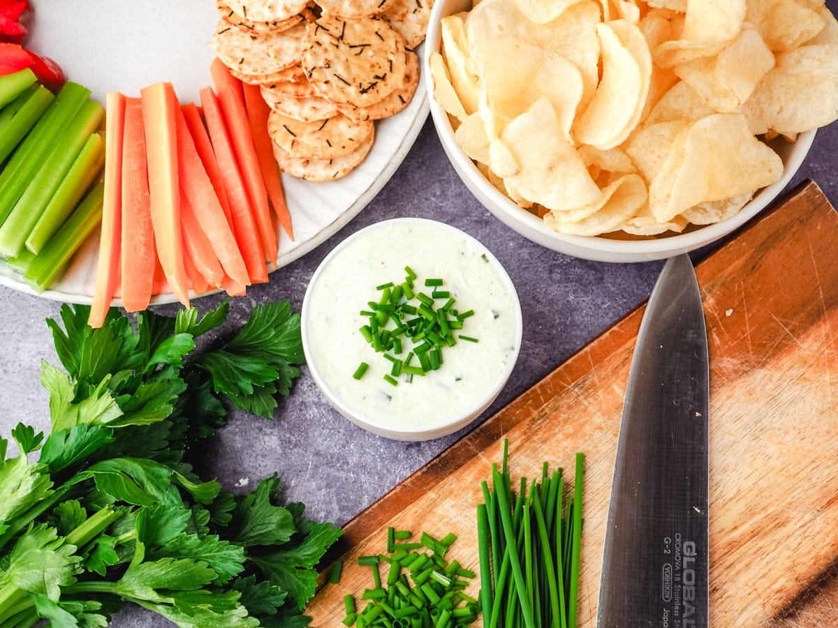 Sour cream dip garnished with fresh chives, with a bowl of chips, plate of crackers and veggie sticks, and chopping board with freshly chopped chives.
