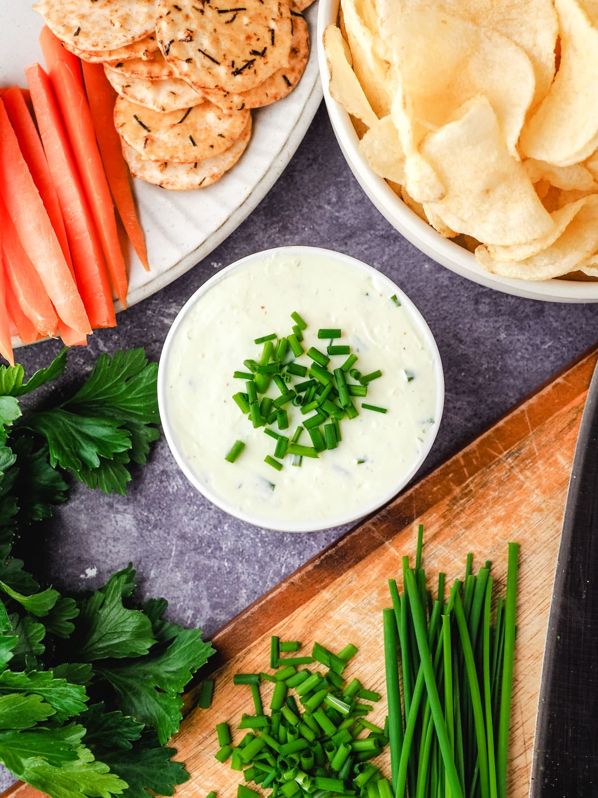 Sour cream dip garnished with fresh chives, with a bowl of chips, plate of crackers and carrot sticks, and chopping board with freshly chopped chives.