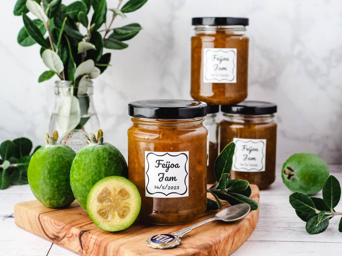 Jar of feijoa jam on a serving board with fresh feijoas and leaves on the side, with a stack of feijoa jam jars in the background.