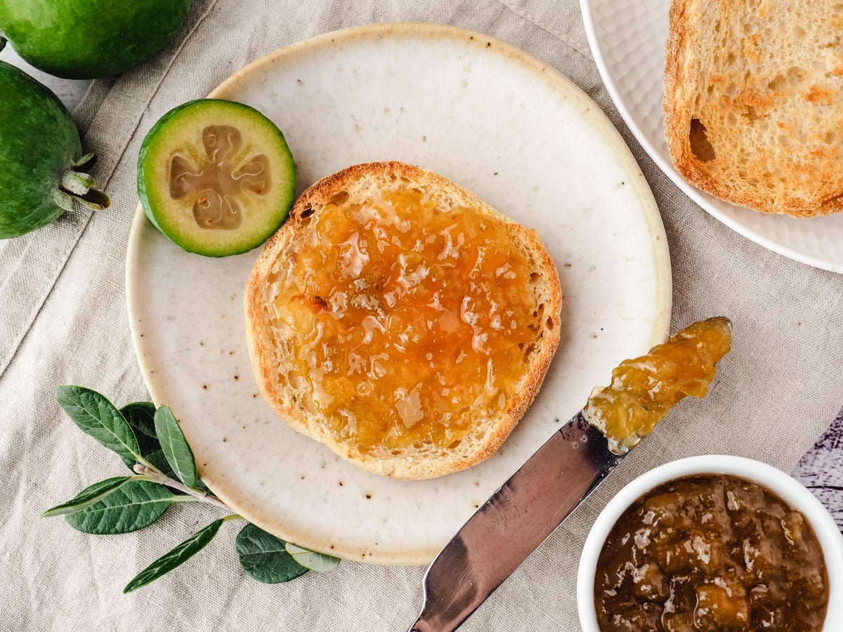 Toasted English muffin spread with feijoa jam, on a plate with a knife and fresh feijoas and pot of jam on the side.