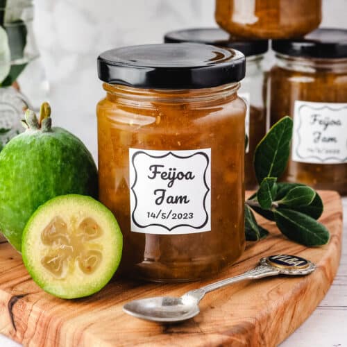Jar of feijoa jam on a serving board with fresh feijoas and leaves on the side, more jars and a plate of toasted muffins in the background.