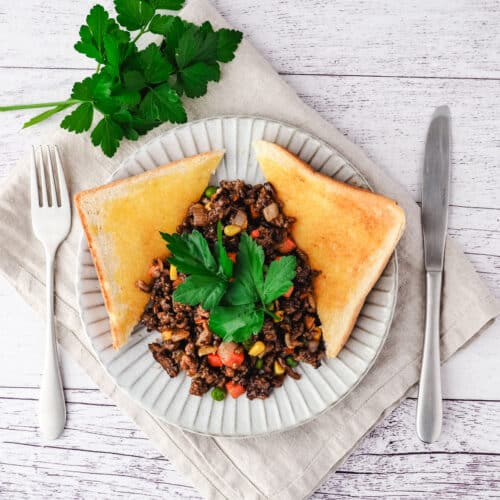 Savory mince on a plate with buttered toast on the side, garnished with flat parsley, with a knife and fork on the side.