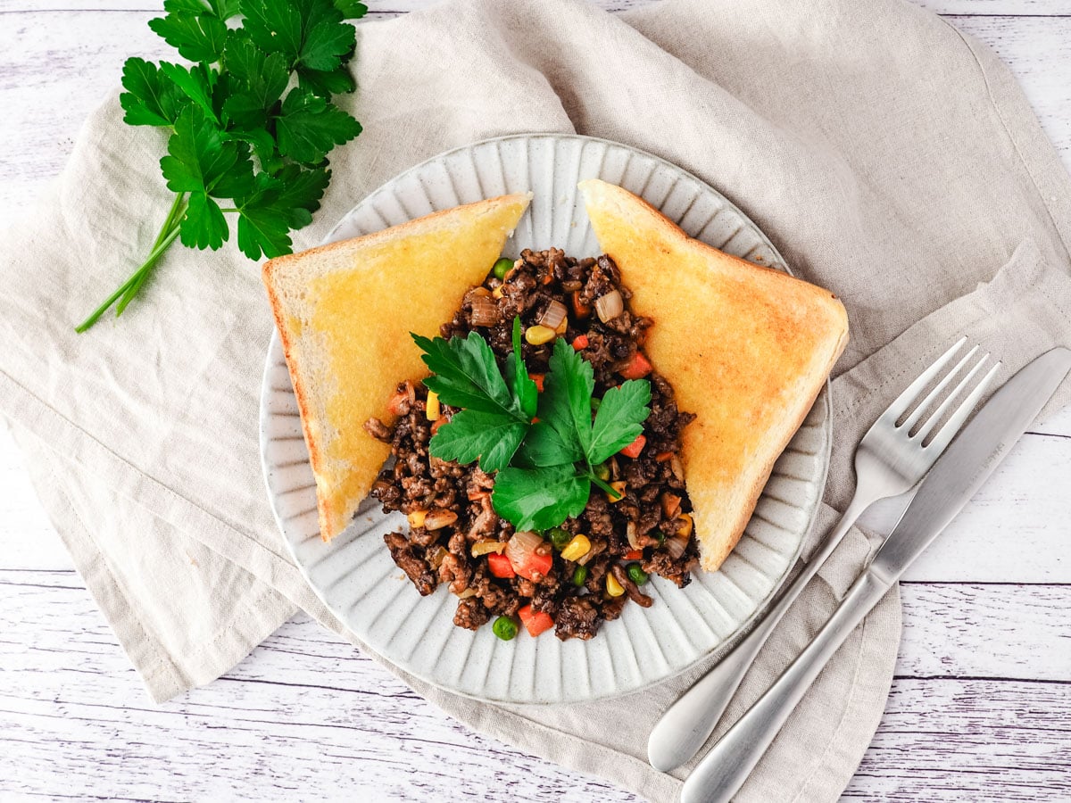 Savory mince on a plate with buttered toast on the side, garnished with flat parsley, with a knife and fork on the side.