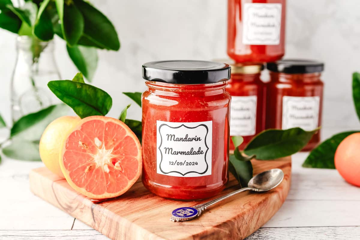 Jar of mandarin marmalade on a serving board with a spoon, with freshly sliced open mandarin and leaves on the side.