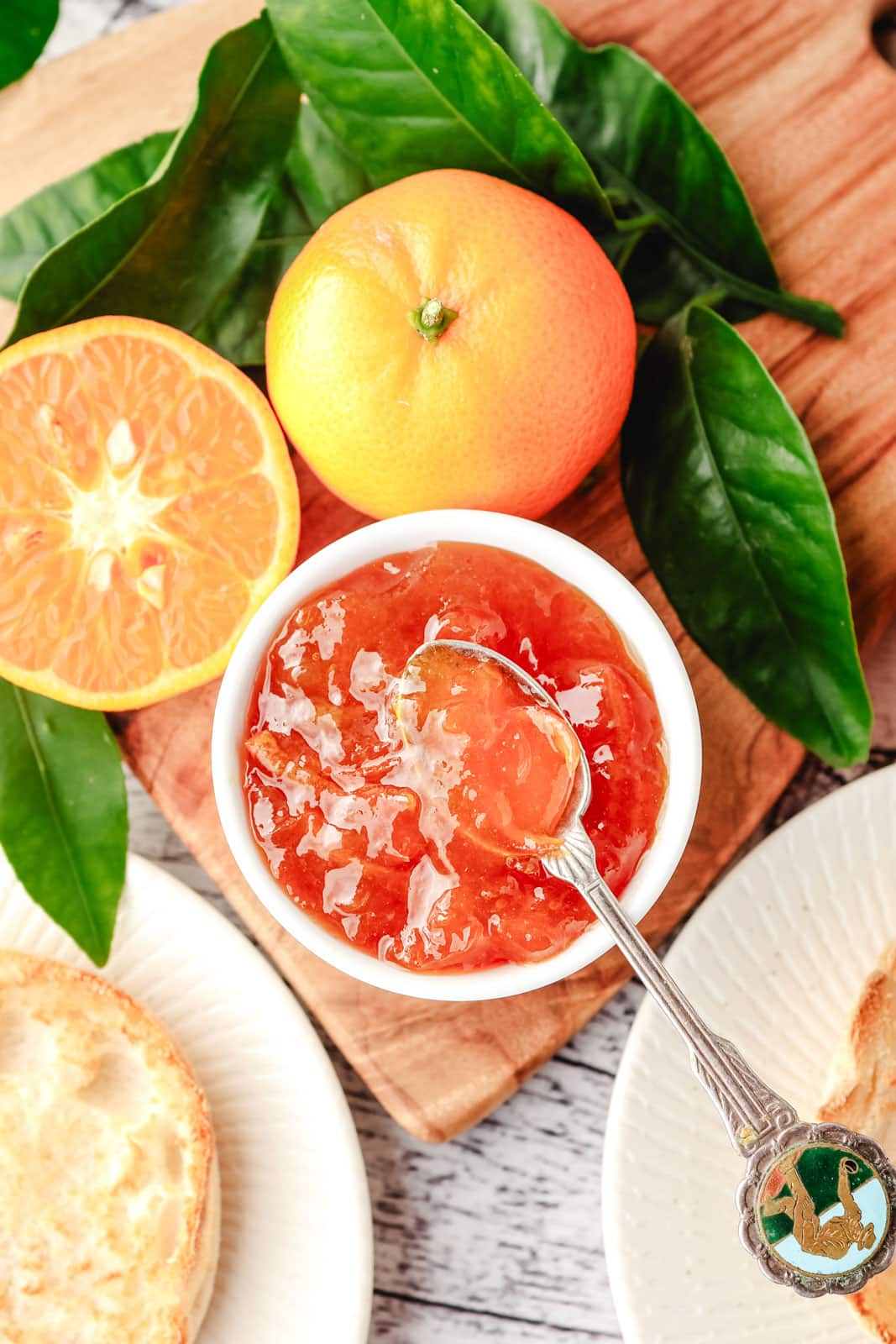 Top down view of mandarin marmalade in a bowl with a spoon, with toasted English muffins and fresh mandarins on the side.