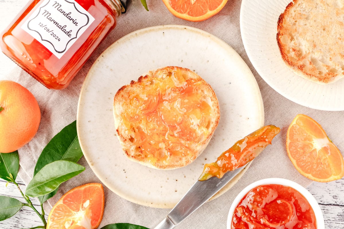 Top down view of mandarin marmalade on a toasted English muffin, on a plate with serving knife and jar of marmalade on the side.